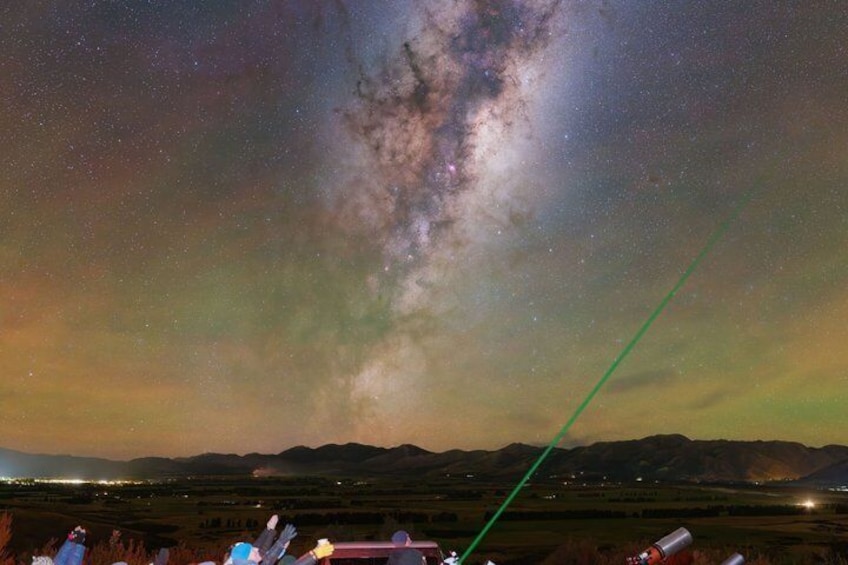 A night sky with the Milky Way, a green laser beam, and a landscape with mountains and lights. 2