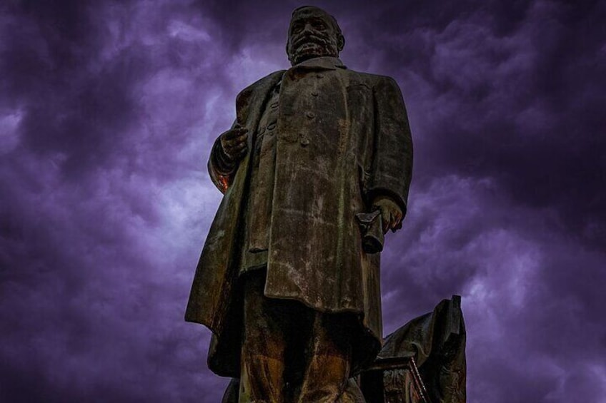 A statue of a man in a long coat and hat, standing against a cloudy sky. 2