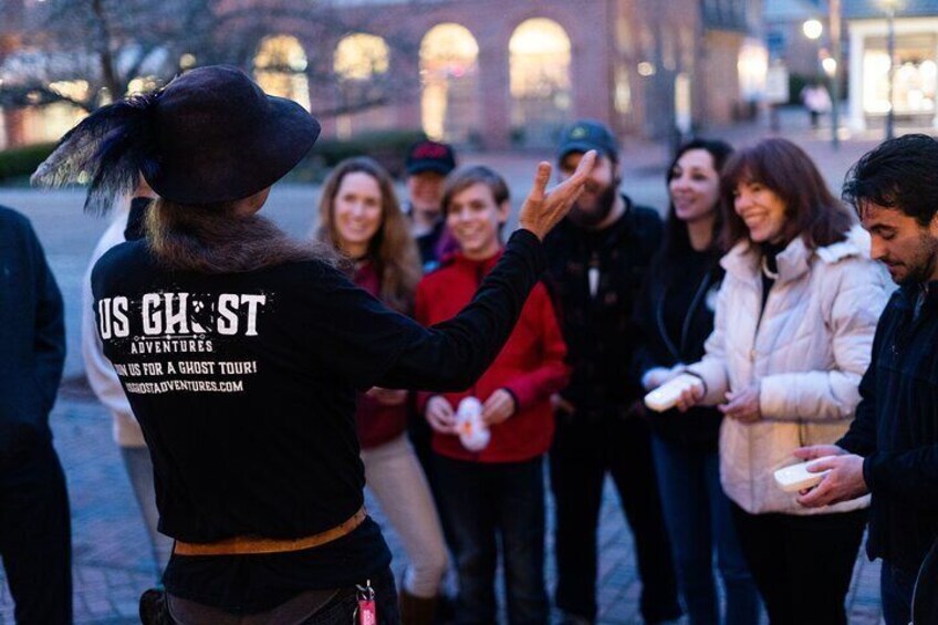 A person in a black hat and jacket with 'US GHOST ADVENTURES' on the back, interacting with a group of people in a public square. 3