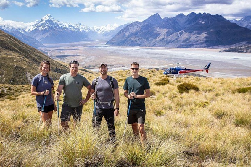 Four people hiking on a grassy field with mountains and a helicopter in the background. 1