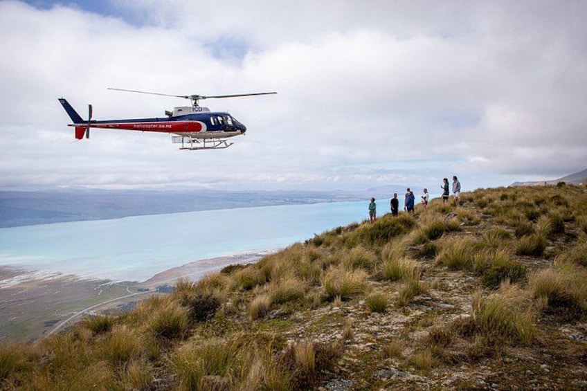 A helicopter flying over a grassy hill with a group of people below. 4