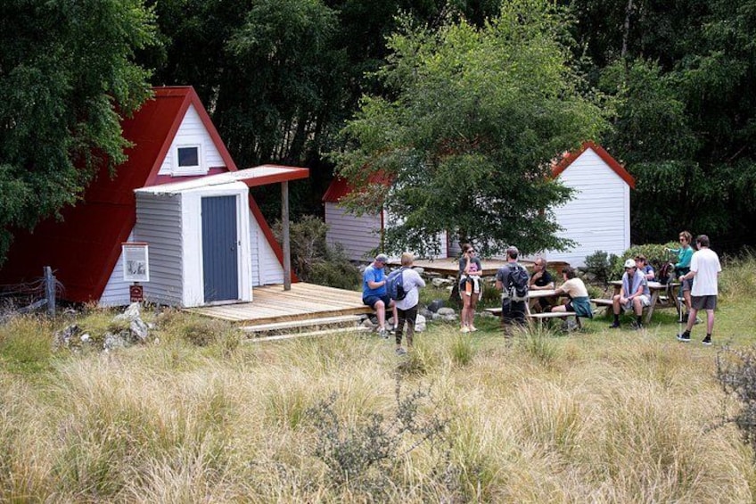 A group of people gathered around a wooden deck in a grassy area with red-roofed cabins in the background. 2