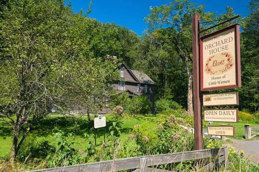 Señal de Orchard House, hogar de Las hermanas pequeñas, con un exuberante paisaje verde y una cerca de madera en primer plano. 1