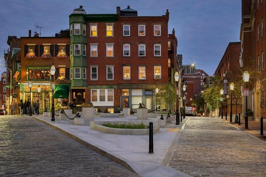 A cobblestone street lined with historic buildings, a public square with a fountain, and street lamps. 2
