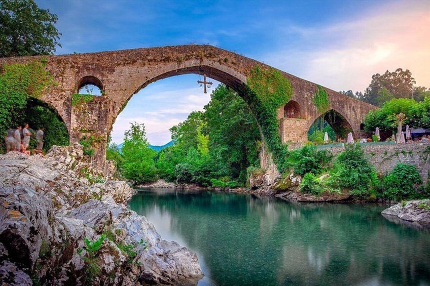 An ancient stone bridge with arches over a calm river, surrounded by lush greenery and rocky terrain. 5