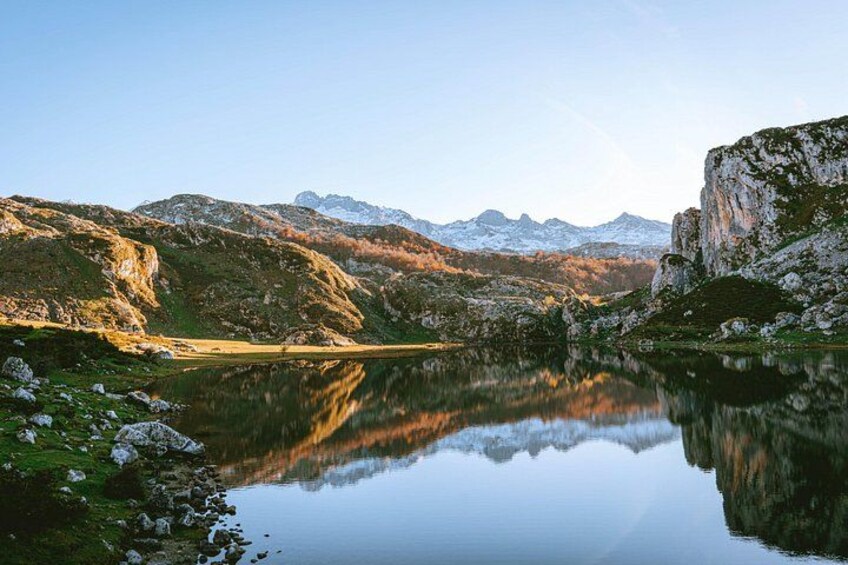 A calm lake reflecting mountains, a clear sky, and a rocky shoreline. 3
