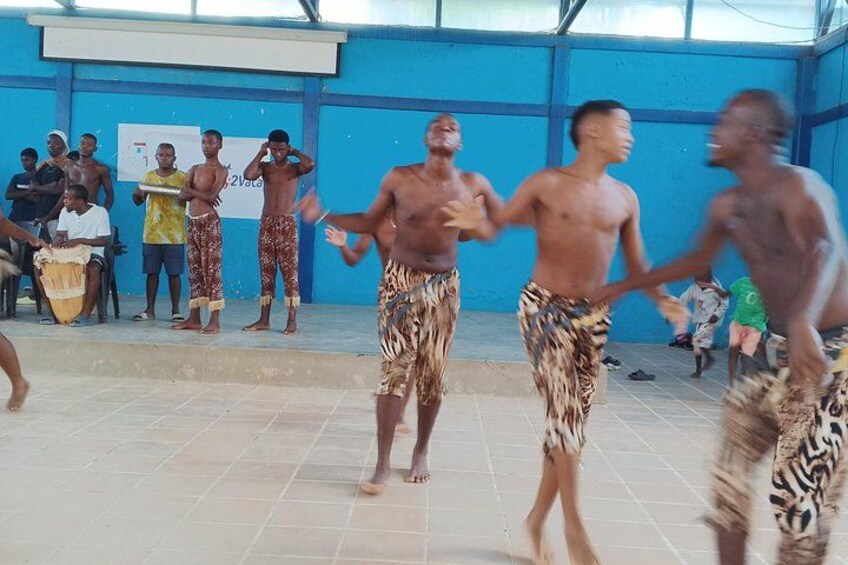 A group of people in patterned shorts dancing in a covered area with a blue wall and a whiteboard in the background. 4
