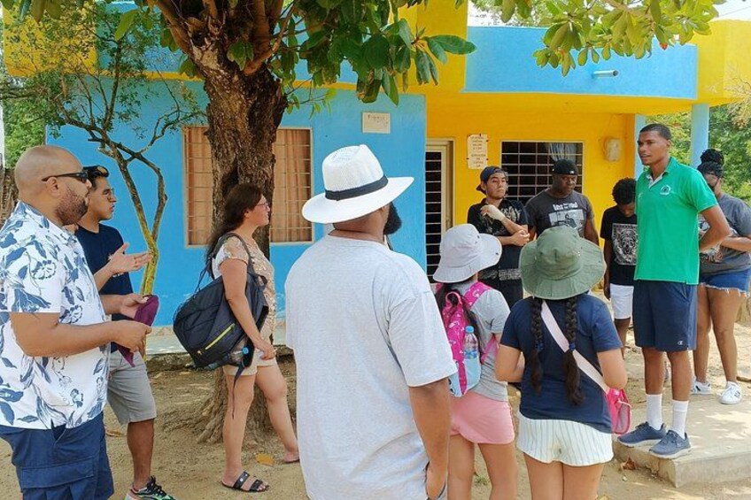 A group of people standing outside a building with a blue and yellow facade, a tree, and a sign on the building. 2