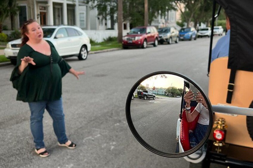Een vrouw gebarend voor een voertuig, een spiegel reflecteert iemand die een foto maakt, en een straat met geparkeerde auto's. 3