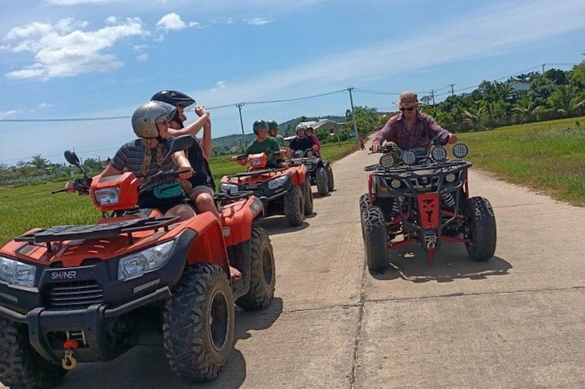 A group of people riding ATVs on a paved road with greenery on the sides. 1