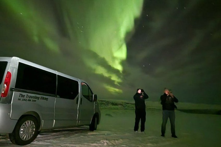 Una camioneta blanca con el texto 'The Traveling Guys', dos personas tomando fotos y la aurora boreal al fondo. 5