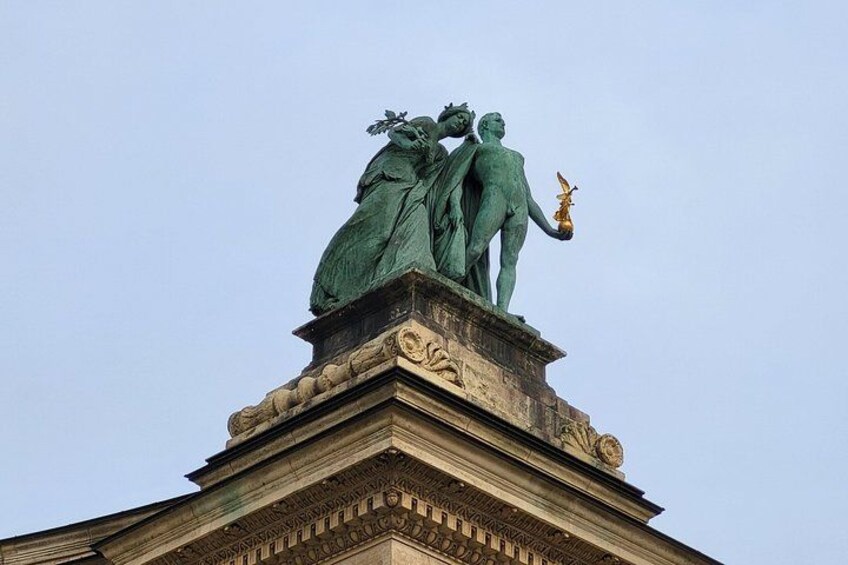 Une statue de bronze, placée au sommet d’un bâtiment de style classique, dont l’architecture présente de nombreux détails complexes. 4