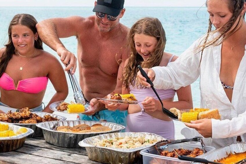 Een groep mensen bij een barbecue aan het strand, met eten in aluminium bakken. 1