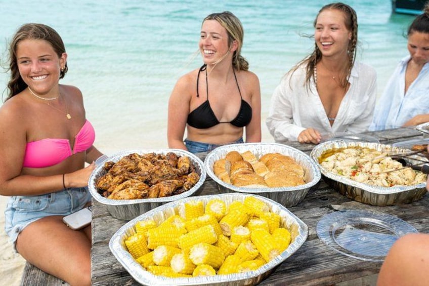 Un gruppo di persone sedute a un tavolo sulla spiaggia, con piatti come pollo alla griglia, pane e pannocchie. 2