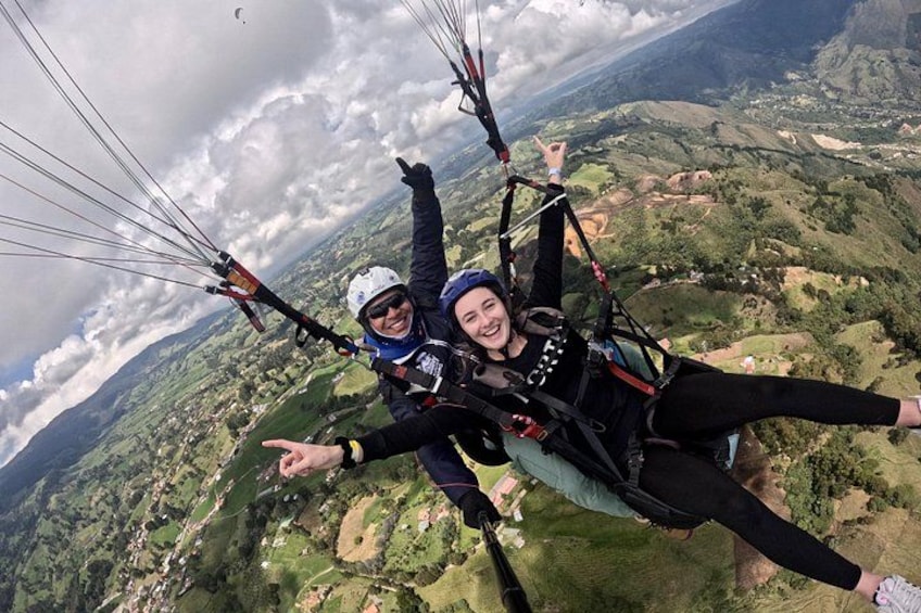 Dos personas hacen parapente sobre un paisaje montañoso con vegetación y pequeños edificios abajo. 3