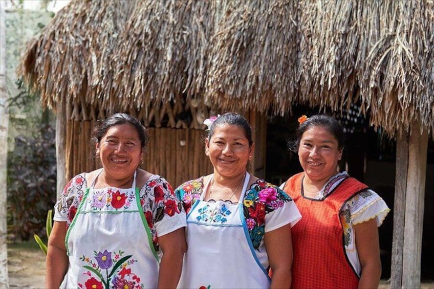 Tres mujeres vestidas con atuendo tradicional frente a una construcción con techo de paja. 4