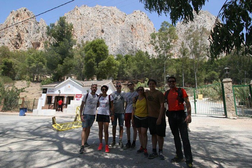 Un grupo de personas de pie en una zona sombreada, con una montaña rocosa de fondo y un pequeño edificio con señalización. 5