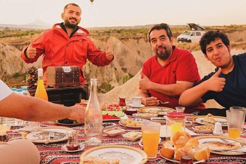 Three people at a table with food and drinks, outdoors with a mountainous landscape in the background. 2