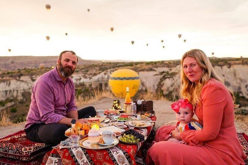 A family picnic with a table set with food and drinks, a child with a red bow, and hot air balloons in the background. 5