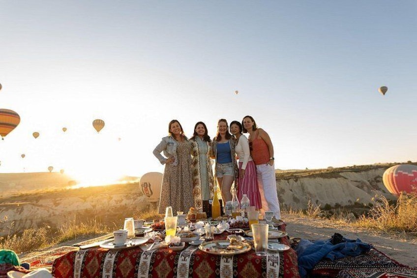 A group of people standing around a table with a meal set outdoors, with hot air balloons in the background. 4