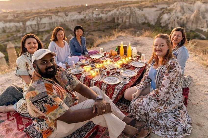A group of people sitting on a blanket outdoors with a table set for a meal. 3