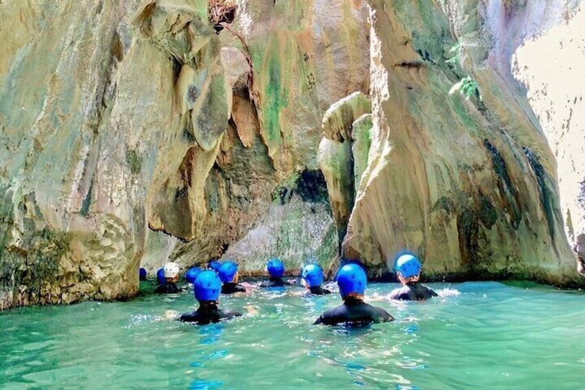 People in blue helmets swimming in a turquoise pool with rocky cliffs in the background. 1