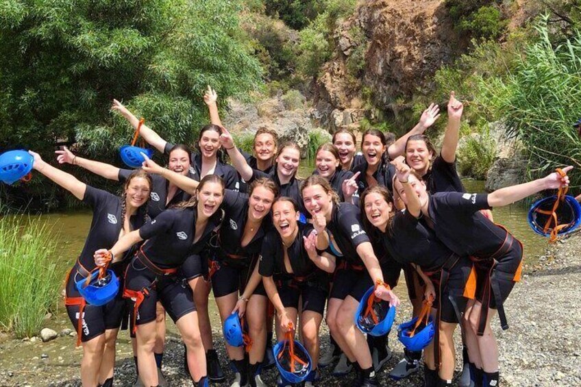 A group of people in black wetsuits with blue helmets and life vests, standing by a river. 2