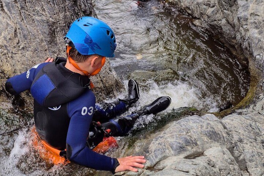 A person in a blue helmet and wetsuit sitting on a rock by a flowing river. 3