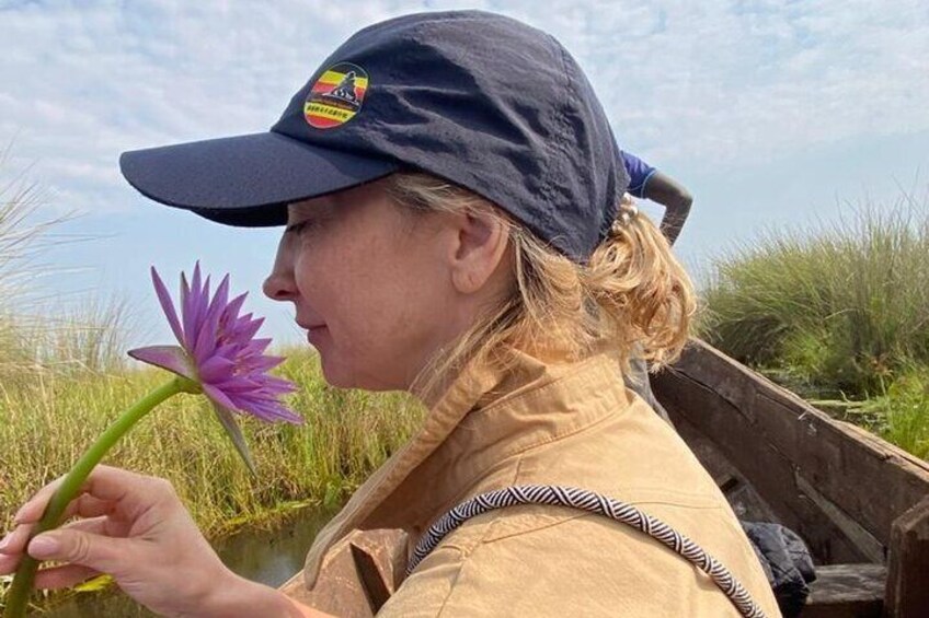 Eine Person hält eine violette Blume neben einem Gewässer, im Hintergrund hohes Gras. 5