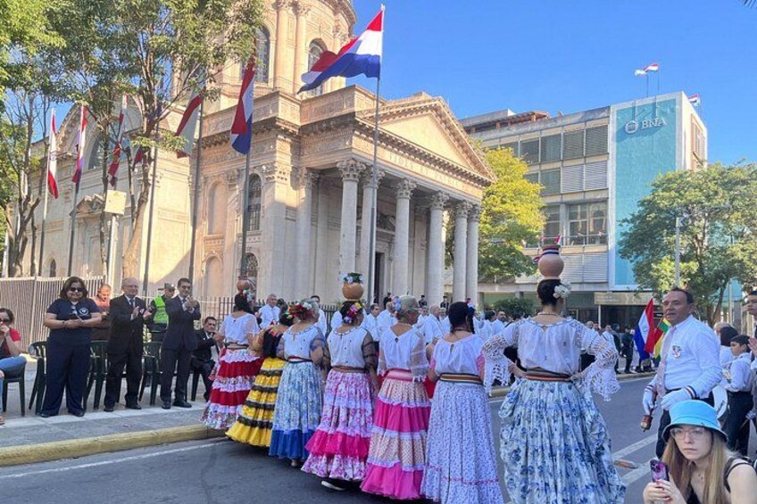 A group of people in traditional attire, a building with columns, and flags. 5