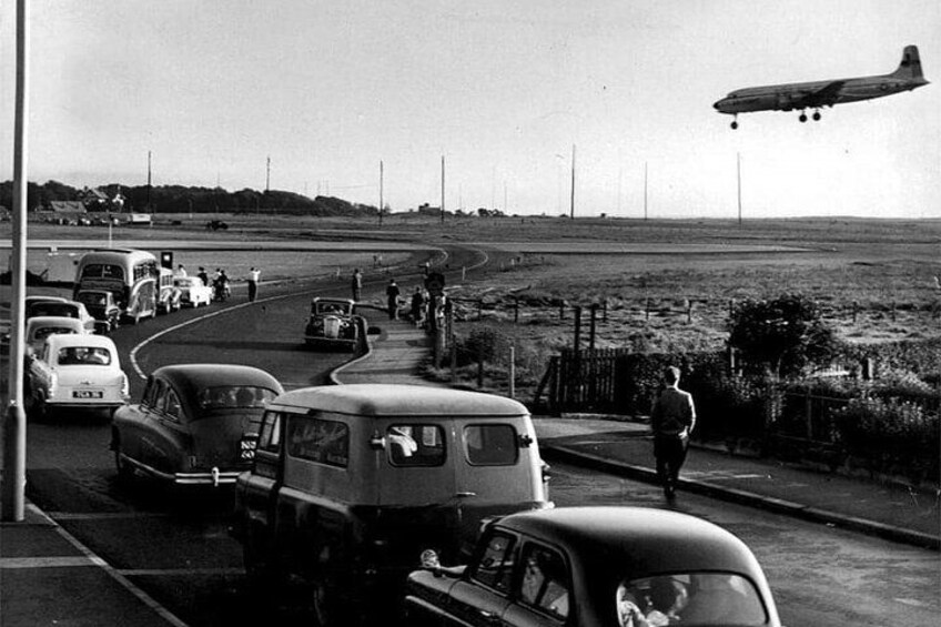 Vintage cars parked on a road with a large airplane landing nearby. 4