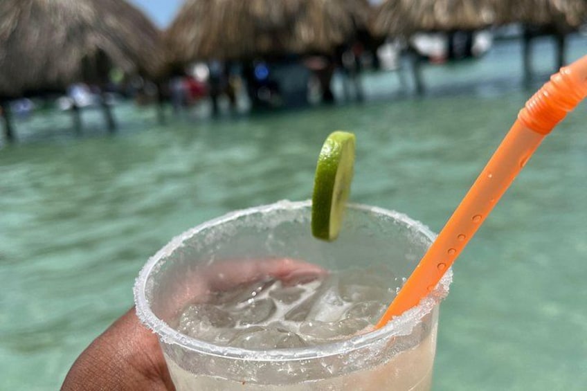 A clear glass with a lime wedge and an orange straw, held over a body of water with thatched structures in the background. 4