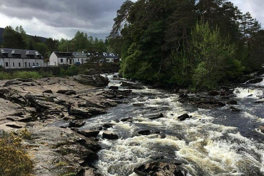 Une rivière aux eaux tumultueuses, bordée de rochers et d’arbres, avec des maisons qui se dessinent à l’arrière-plan. 2