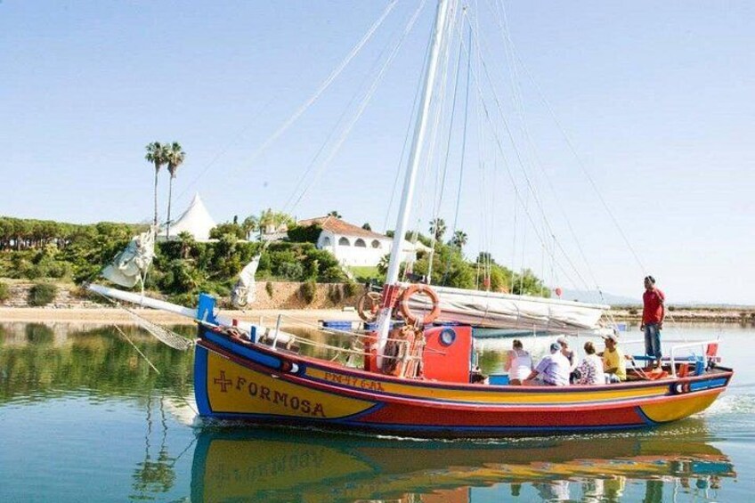 Un barco colorido llamado 'FORMOSA' amarrado en una orilla tranquila, con cielo despejado y un edificio al fondo. 1