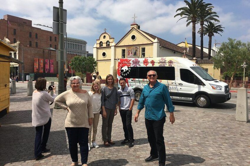 Een groep mensen staat op een geplaveid straatje, met een kerk en een witte bus op de achtergrond. 3