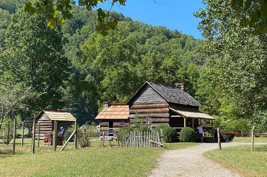 A rustic log cabin with a gravel path leading to it, surrounded by lush greenery and a forested hill in the background. 1