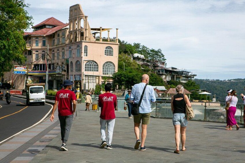 A pedestrian walkway with people walking, a road with a white vehicle, and a large building with a red roof. 4