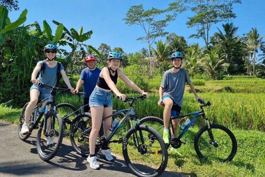 Four people on bicycles in a rural setting with lush greenery and palm trees. 1
