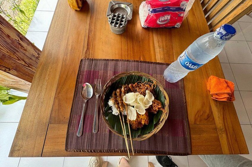A wooden table with a woven placemat, a bowl of food, chopsticks, a spoon, a bottle of water, and a packet of Jolly. 3