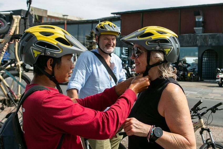 Three people wearing helmets, one adjusting another's helmet, with bicycles and a building in the background. 5