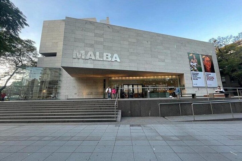 A modern building with the name 'MALBA' on its facade, surrounded by a paved area with steps leading up to the entrance. 2