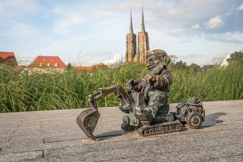Eine bronzene Statue eines Mannes auf einem Motorrad mit einer Schaufel, auf einer Betonfläche, im Hintergrund eine Kirche und Gebäude. 4