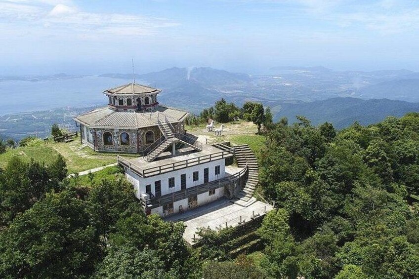 Ein historisches Gebäude mit einem Turm und einem niedrigeren Anbau, umgeben von üppiger Vegetation und Bergen im Hintergrund. 3