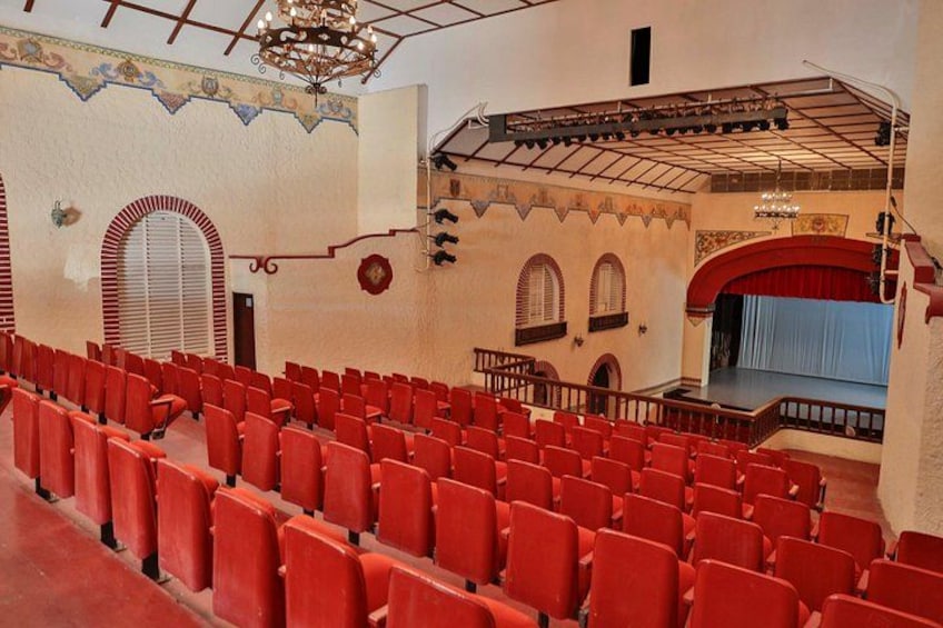An indoor auditorium with red seats, a stage with a blue curtain, and ornate wall decorations. 3