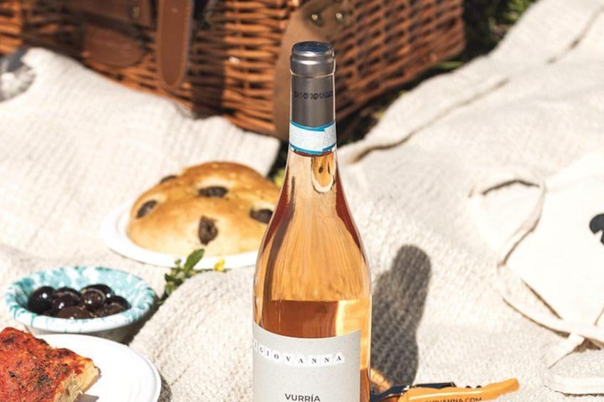 A bottle of Vurria wine, a wicker basket, a plate with a pastry, and a bowl of olives on a picnic blanket. 3
