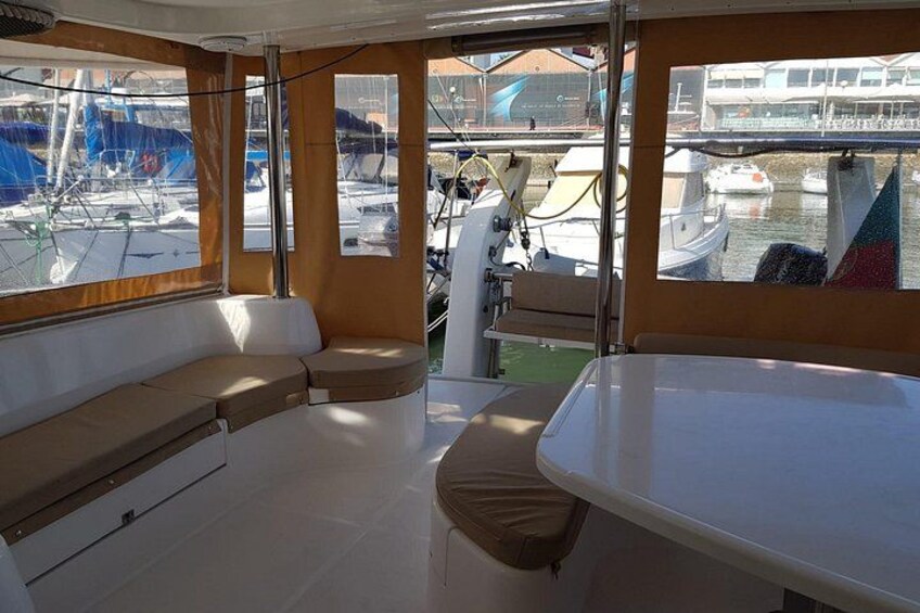 Schiffsinnenraum mit gepolsterten Sitzen, einem Tisch und Fenstern mit Blick auf eine Marina und weitere Boote. 5