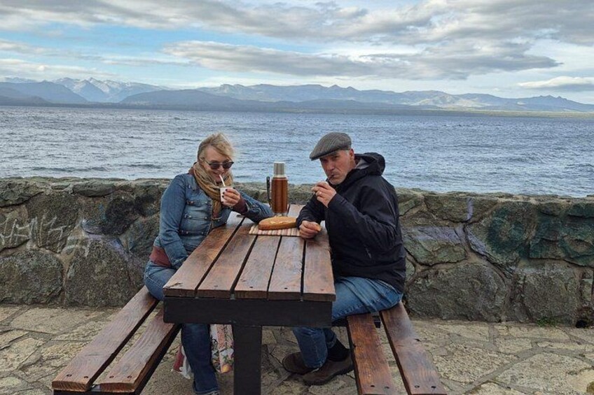 Deux personnes sont assises à une table en bois, près d’une côte rocheuse, avec une étendue d’eau et des montagnes en arrière-plan. 3