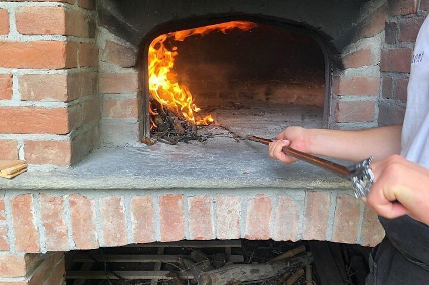 A brick oven with flames inside, a person using a tool to manage the fire, and a partially visible brick wall. 1