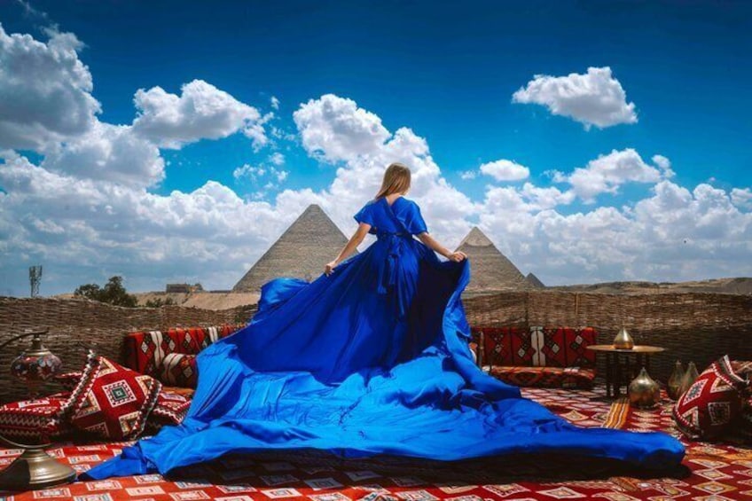 A person in a blue dress standing on a patterned carpet with a pyramid in the background. 1