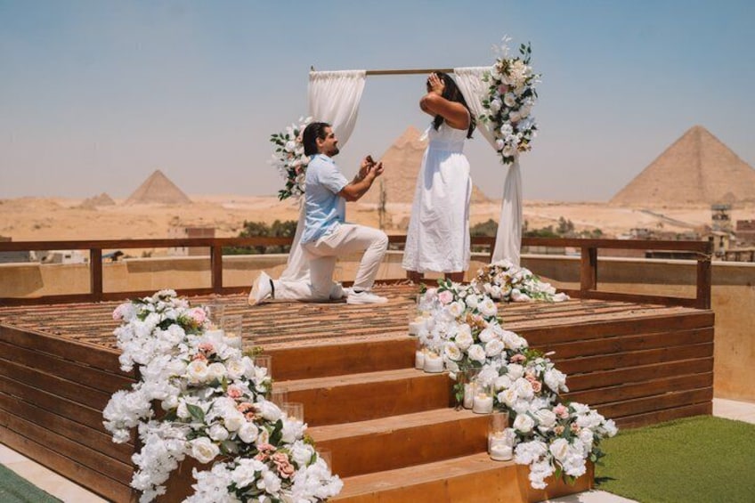 A man kneeling on a wooden platform with a woman standing in front of a backdrop with flowers, with pyramids in the background. 4